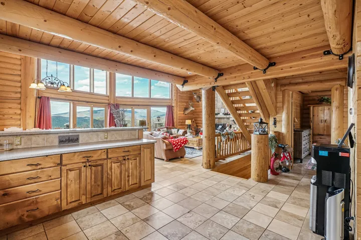 Great room featuring expansive window walls, natural wood log construction, exposed beam ceilings, a built-in kitchen peninsula with a light-toned countertop, and tiled flooring