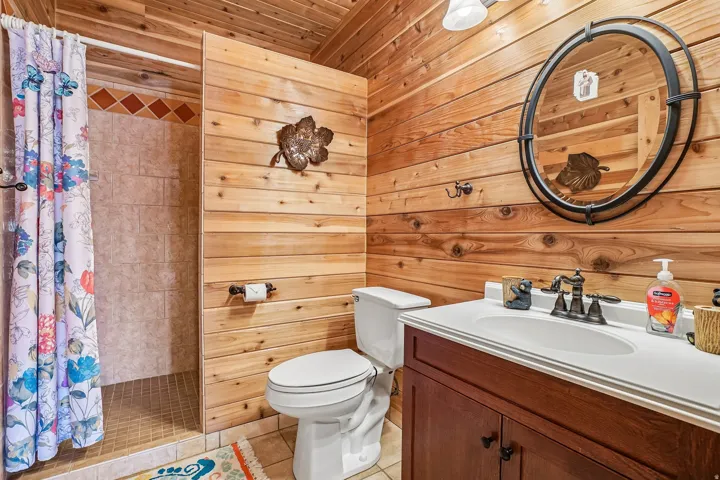 Bathroom featuring wood-paneled walls and ceiling, a walk-in shower with tile surround, a single vanity with a white countertop and dark wood-finish cabinetry, and a round wall-mounted mirror