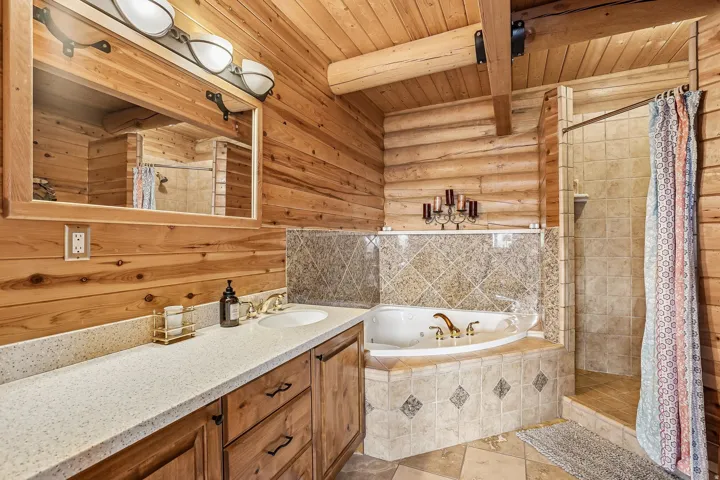 Bathroom featuring wood-paneled walls, a double vanity with an integrated sink, a jetted corner tub with tiled surround, a tiled shower, and exposed wood ceiling beams