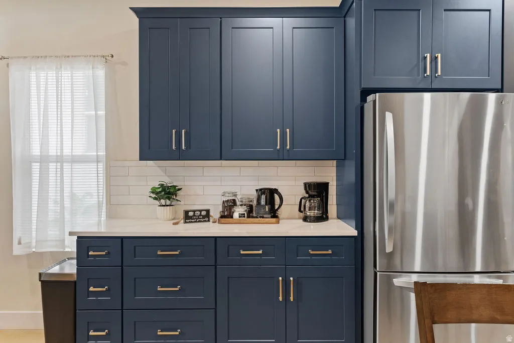 Kitchenette featuring blue shaker-style cabinetry, white subway tile backsplash, light-toned countertop, and stainless steel refrigerator