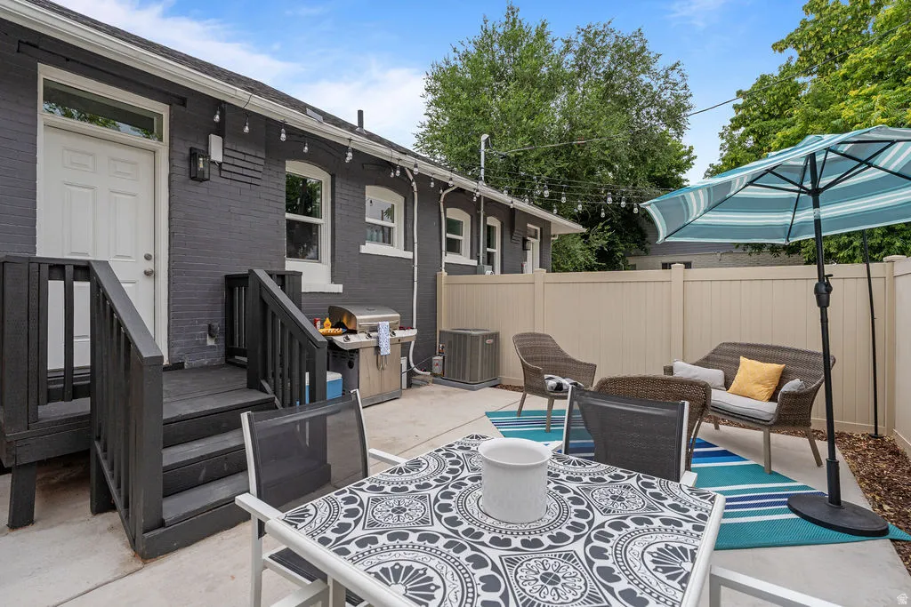 Concrete patio featuring a dark-painted brick exterior with white trim windows, a dark wood deck with steps, string lighting, and a neutral-toned privacy fence