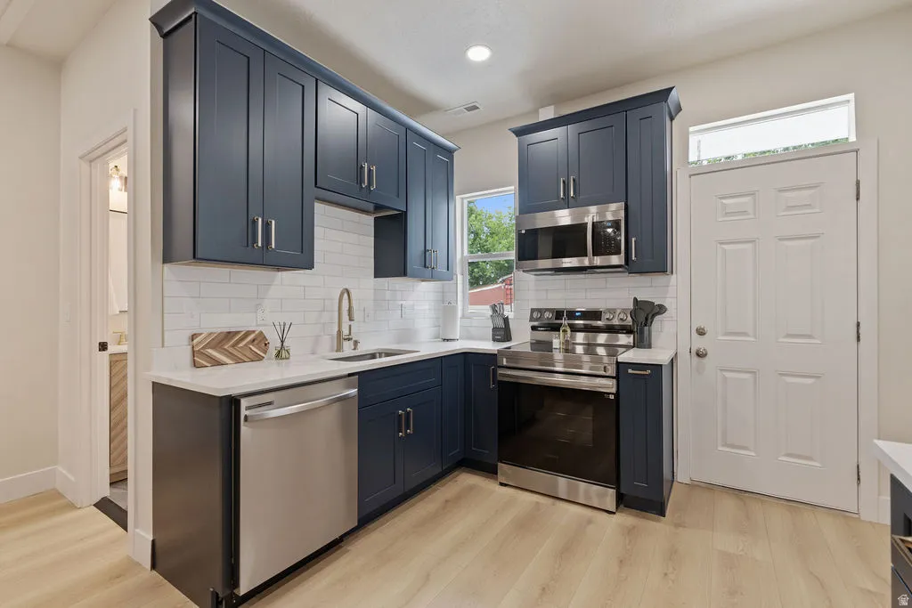 Modern kitchen featuring dark blue cabinetry, stainless steel appliances, white subway tile backsplash, and light wood-finish flooring