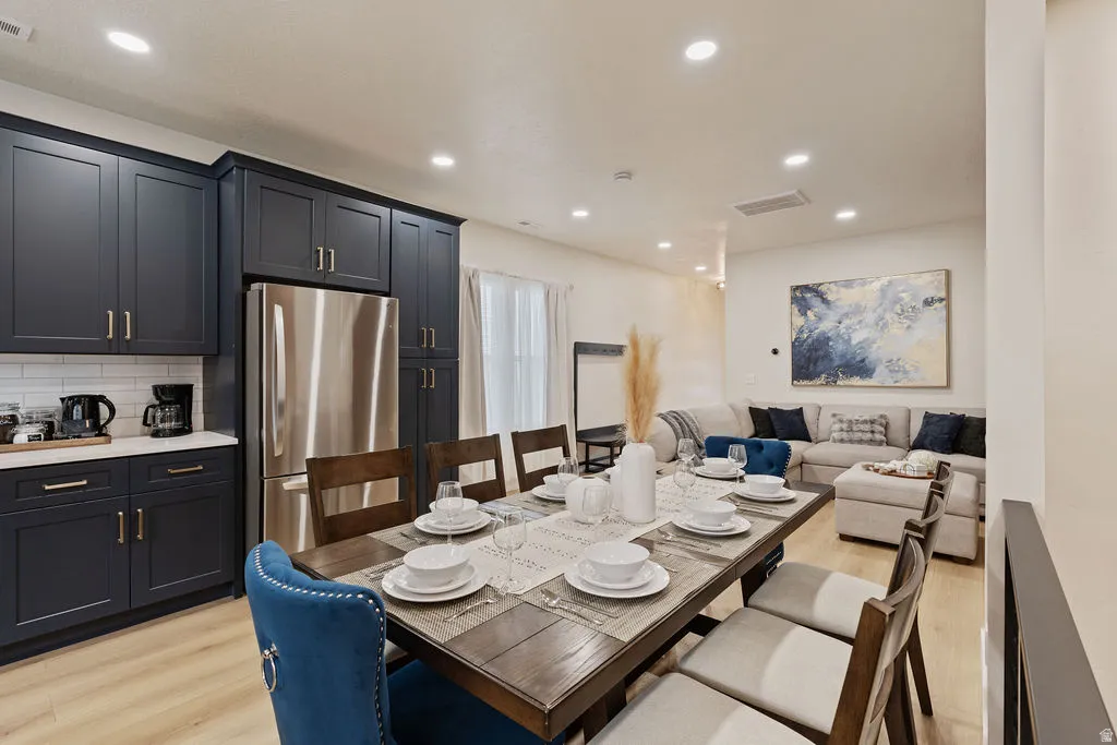Open-concept living area featuring light wood-finish flooring, recessed lighting, and a kitchen with dark cabinetry, stainless steel refrigerator, and white subway tile backsplash