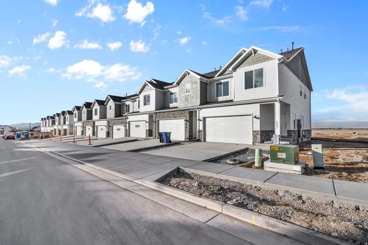 Row of newly constructed townhomes featuring white siding, dark shingle roofs, stone accents, attached garages, and a paved driveway