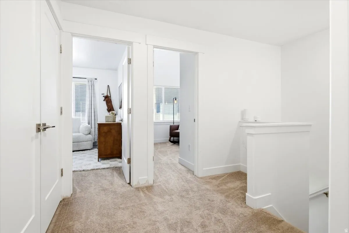 Carpeted hallway with white walls and trim, featuring two doorways leading to additional rooms, a white banister, and a white door with brushed nickel hardware