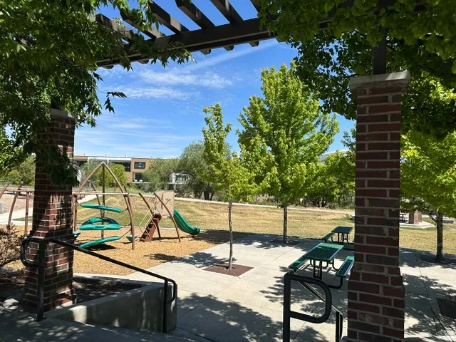 Community park featuring a play structure, picnic tables, open lawn space, mature trees, and a shaded pergola structure with brick pillars