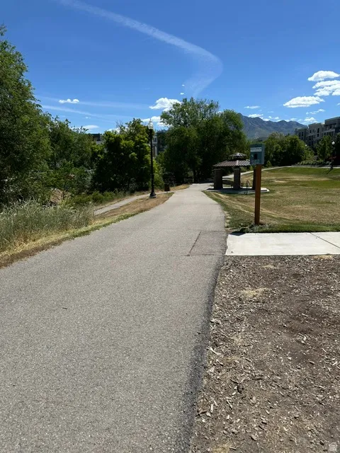 Paved pathway bordered by green spaces, featuring mature trees, a covered gazebo, and distant mountains