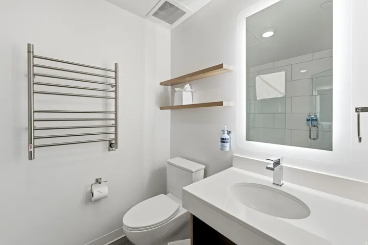 Modern bathroom featuring a vanity with an integrated sink, illuminated mirror, wall-mounted towel warmer, and floating wood-finish shelving