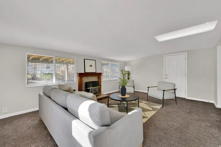 Spacious living area featuring light gray walls, brown carpet flooring, multiple windows with horizontal blinds, a fireplace with a wood mantel, and a white paneled entry door