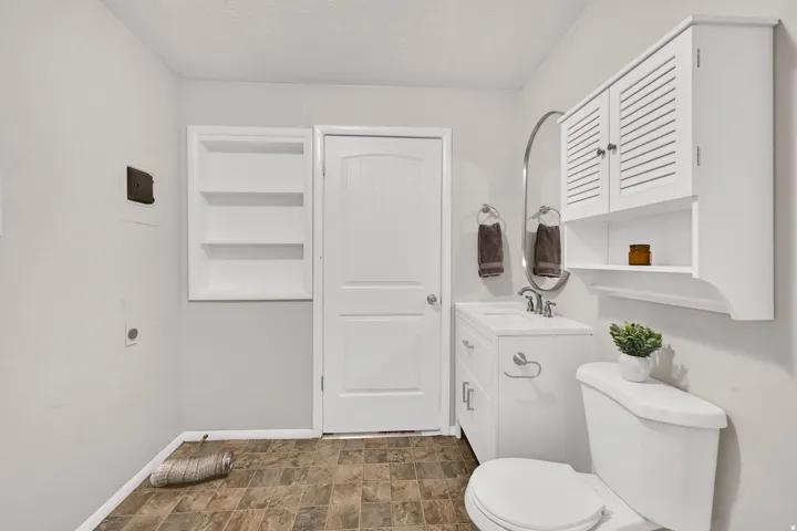 Compact bathroom featuring a white vanity with integrated sink, oval mirror, and wall-mounted shelving above the toilet