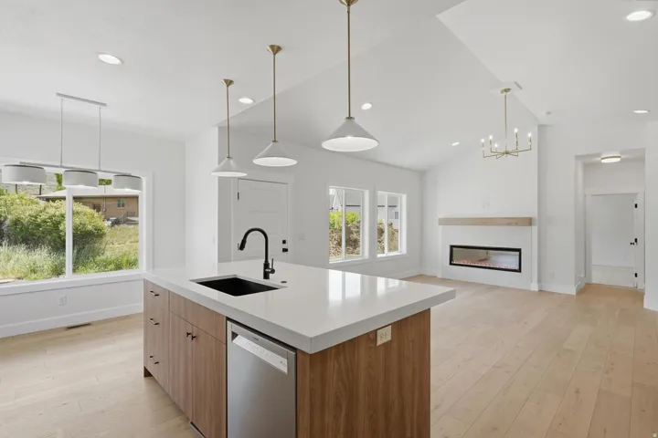 Open-concept living area featuring a kitchen island with integrated sink and dishwasher, light wood-finish flooring, a contemporary fireplace with a wood mantle, and vaulted ceilings with recessed and pendant lighting