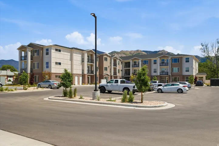 Modern multi-story apartment building featuring balconies with metal railings, a paved parking area, and landscaped islands with young trees