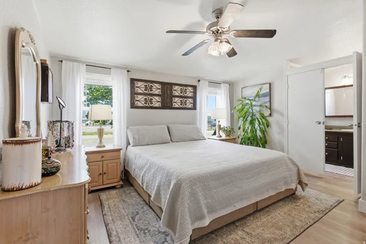 Bedroom with wood-finish flooring, two large windows, a ceiling fan with integrated lighting, and direct access to a bathroom featuring a dark wood vanity and mirror