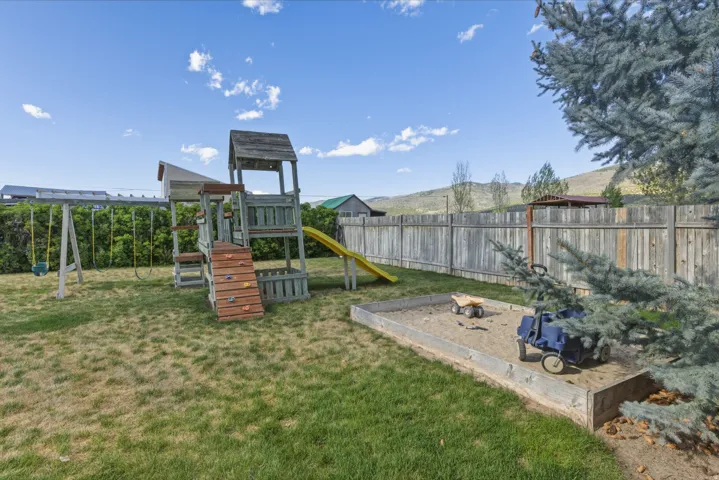 Expansive grassy yard featuring a wooden play structure with slide and swings, a built-in sandbox, and a privacy fence