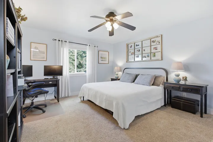 Carpeted bedroom featuring a window with white curtains, a ceiling fan with integrated lighting, and a dark-toned built-in bookcase