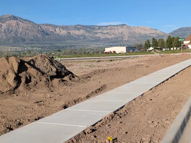 New concrete sidewalk and curb in a developing area, framed by expansive mountain scenery
