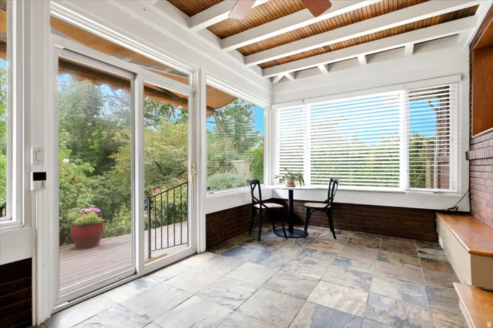 Sunroom featuring exposed wood ceiling beams, a sliding glass door, large windows with horizontal blinds, stone tile flooring, and a built-in wood bench