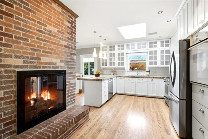 Brick fireplace with hearth, wood-finish flooring, white cabinetry with glass-front uppers, stainless steel appliances, and a kitchen island