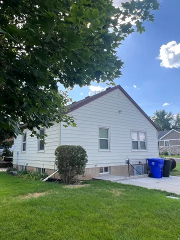 Exterior features include light-colored horizontal siding, a gabled roofline with brown shingles, multiple windows, and a concrete patio