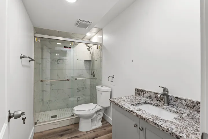 Contemporary bathroom featuring a glass-enclosed shower with white marble-look tile, wood-finish flooring, a white porcelain toilet, and a vanity with a stone countertop and undermount sink