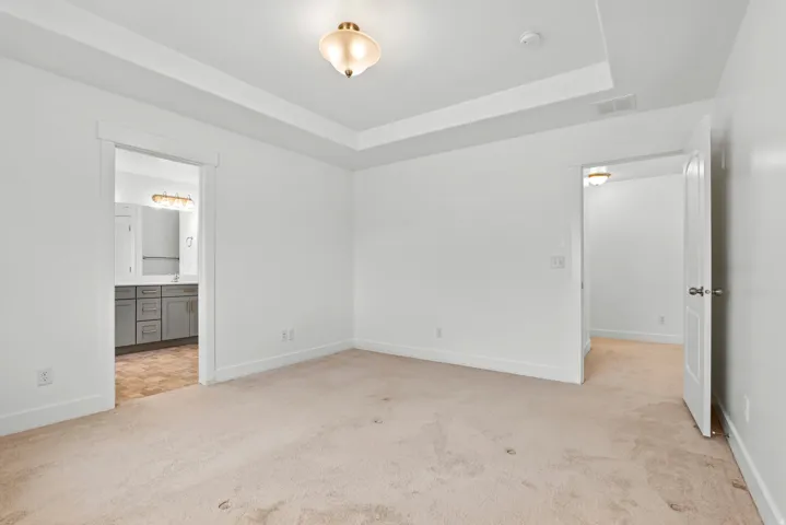 Carpeted room featuring a tray ceiling with a semi-flush mount light fixture, white trim, and a doorway opening to a bathroom with gray cabinetry and a vanity mirror