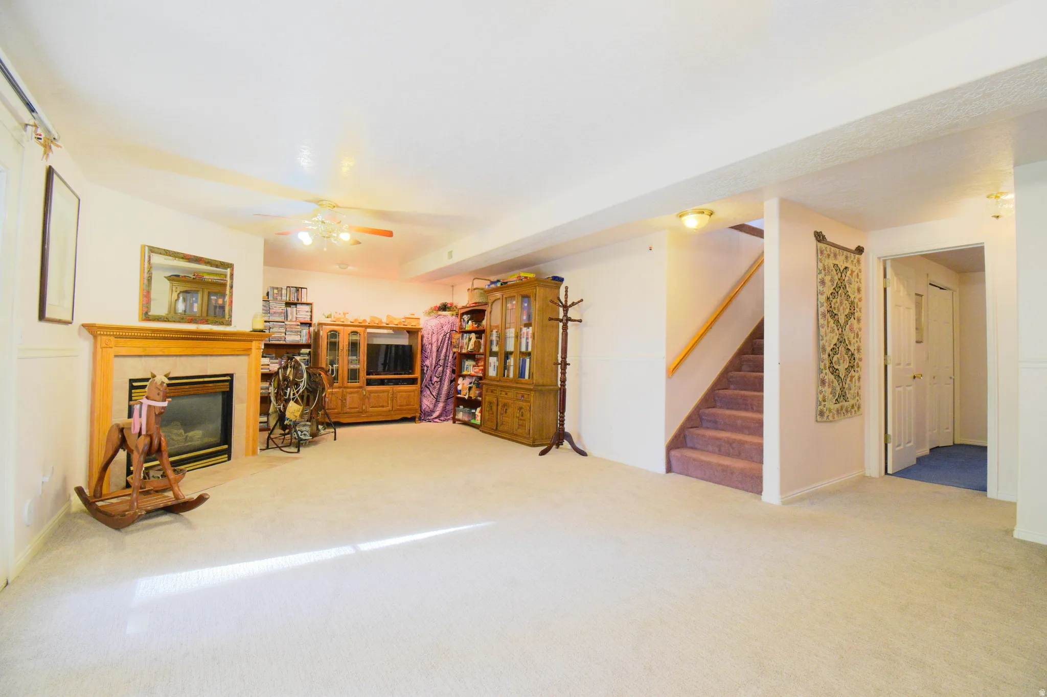 Carpeted recreation room featuring a corner fireplace with wood surround, white walls, built-in ceiling lighting, and a staircase with a wood handrail