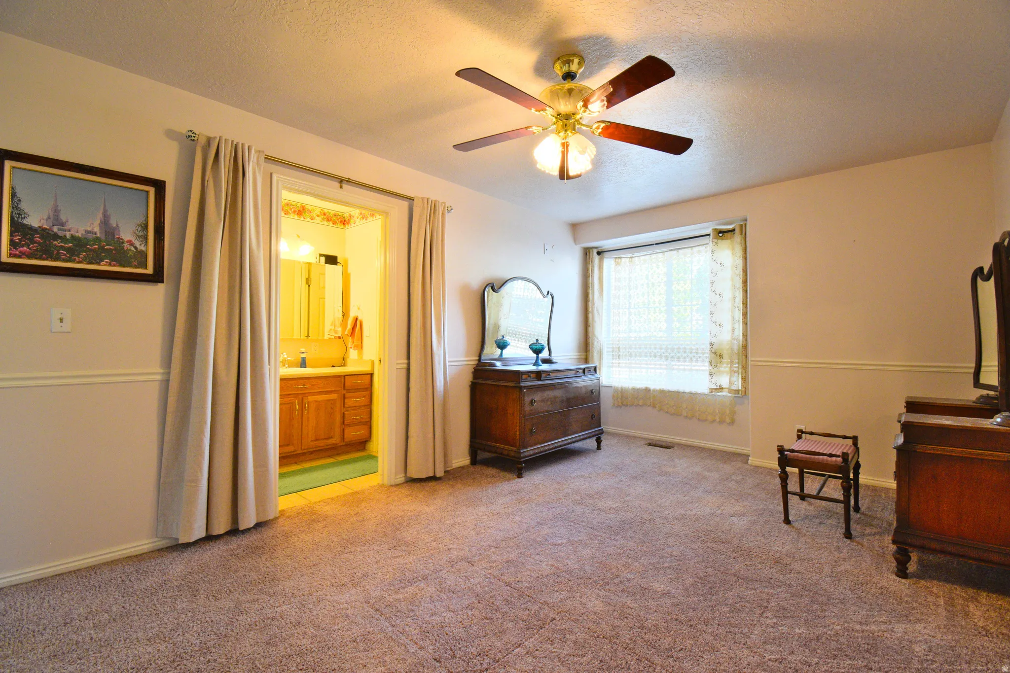 Spacious carpeted room featuring a ceiling fan with integrated lighting, a window with curtains, and direct access to an attached bathroom