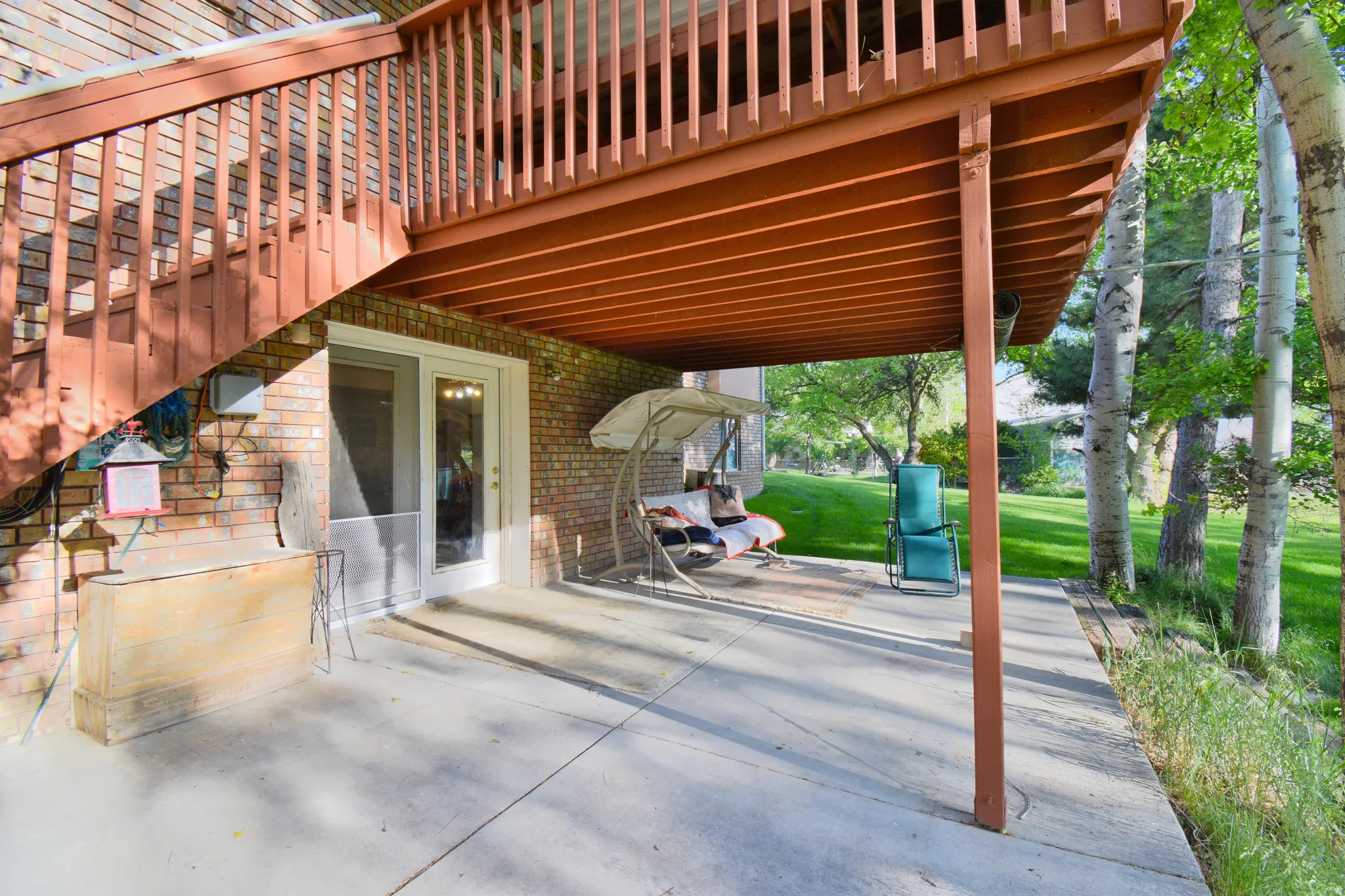 Covered concrete patio with direct outdoor access via a white-framed glass door, featuring a brick exterior wall and a wooden deck overhead