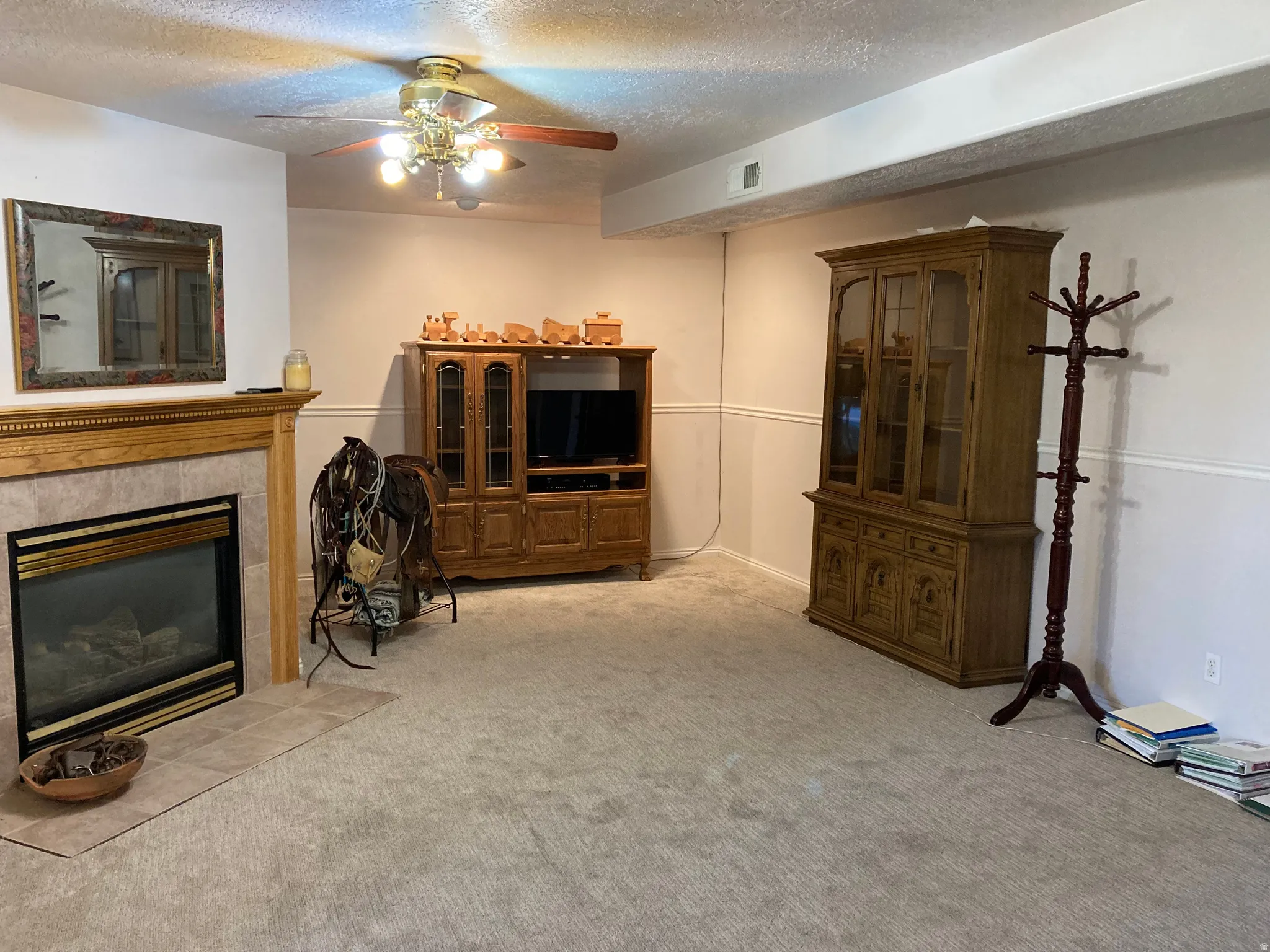 Spacious carpeted room featuring a ceiling fan with light fixture, a tile-surrounded fireplace with a wood mantle, and wainscoting