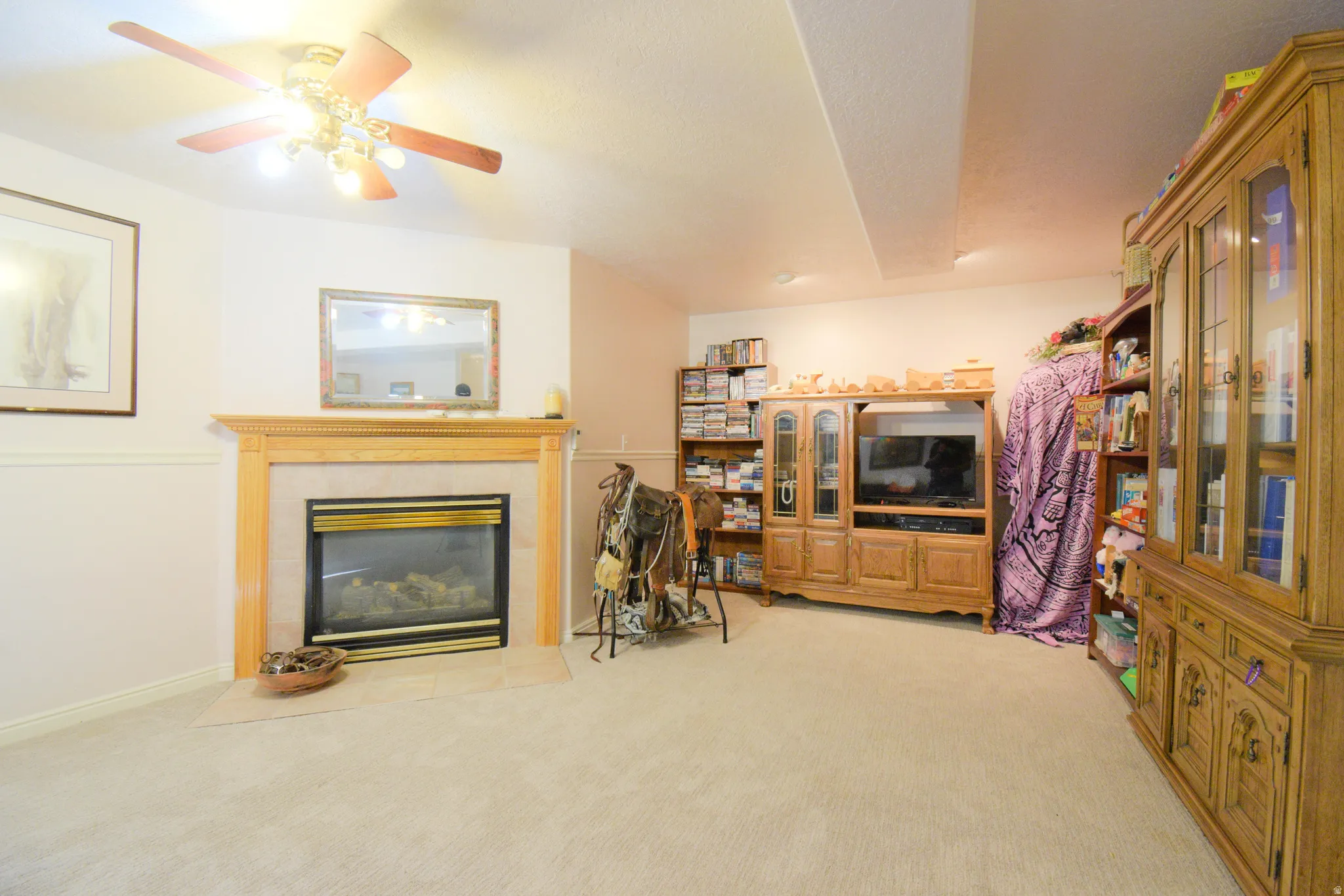 Recessed gas fireplace with a wood mantel and light-colored tile surround, accompanied by plush light-colored carpeting and a ceiling fan with integrated lighting