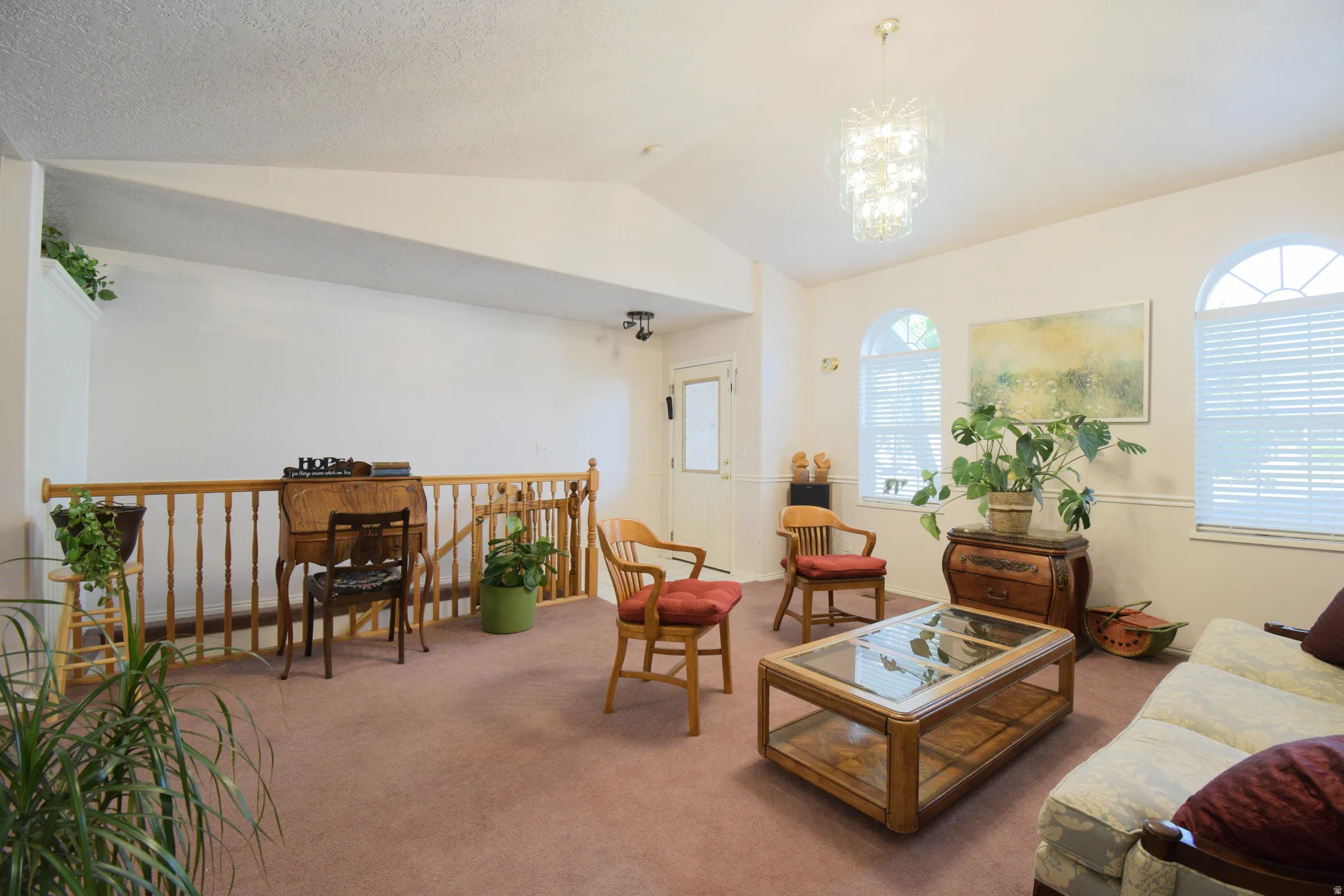 Carpeted living area featuring a wood-finish railing, arched windows with blinds, a multi-tier chandelier, and vaulted ceilings