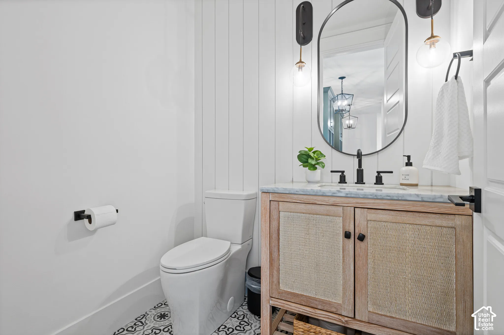 Modern powder room featuring a white shiplap accent wall, patterned floor tile, a vanity with a light wood finish and woven cane doors, a light-toned countertop, and matte black fixtures