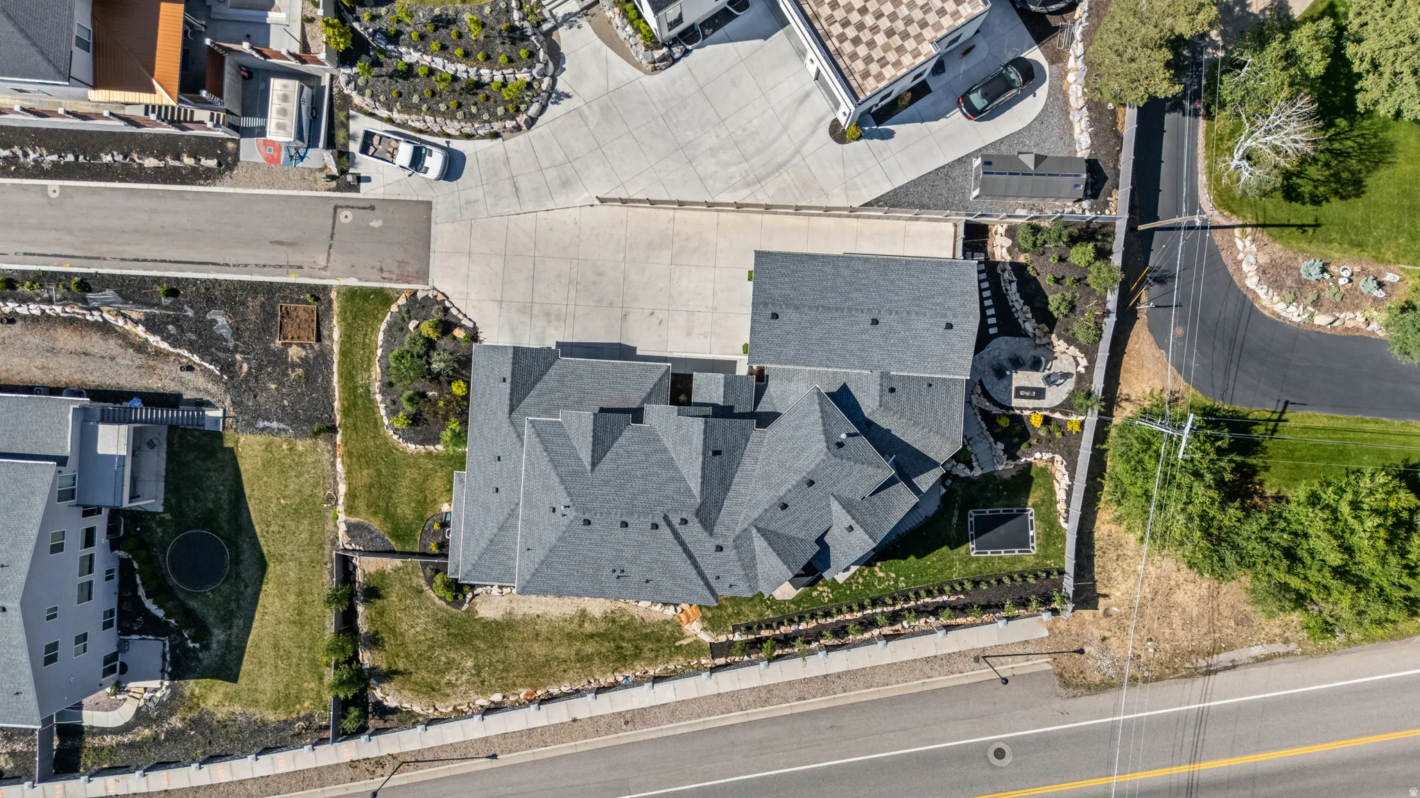 Contemporary residence featuring a multi-gabled gray shingle roof, extensive concrete driveway, landscaped grounds with established plantings, and a stone retaining wall