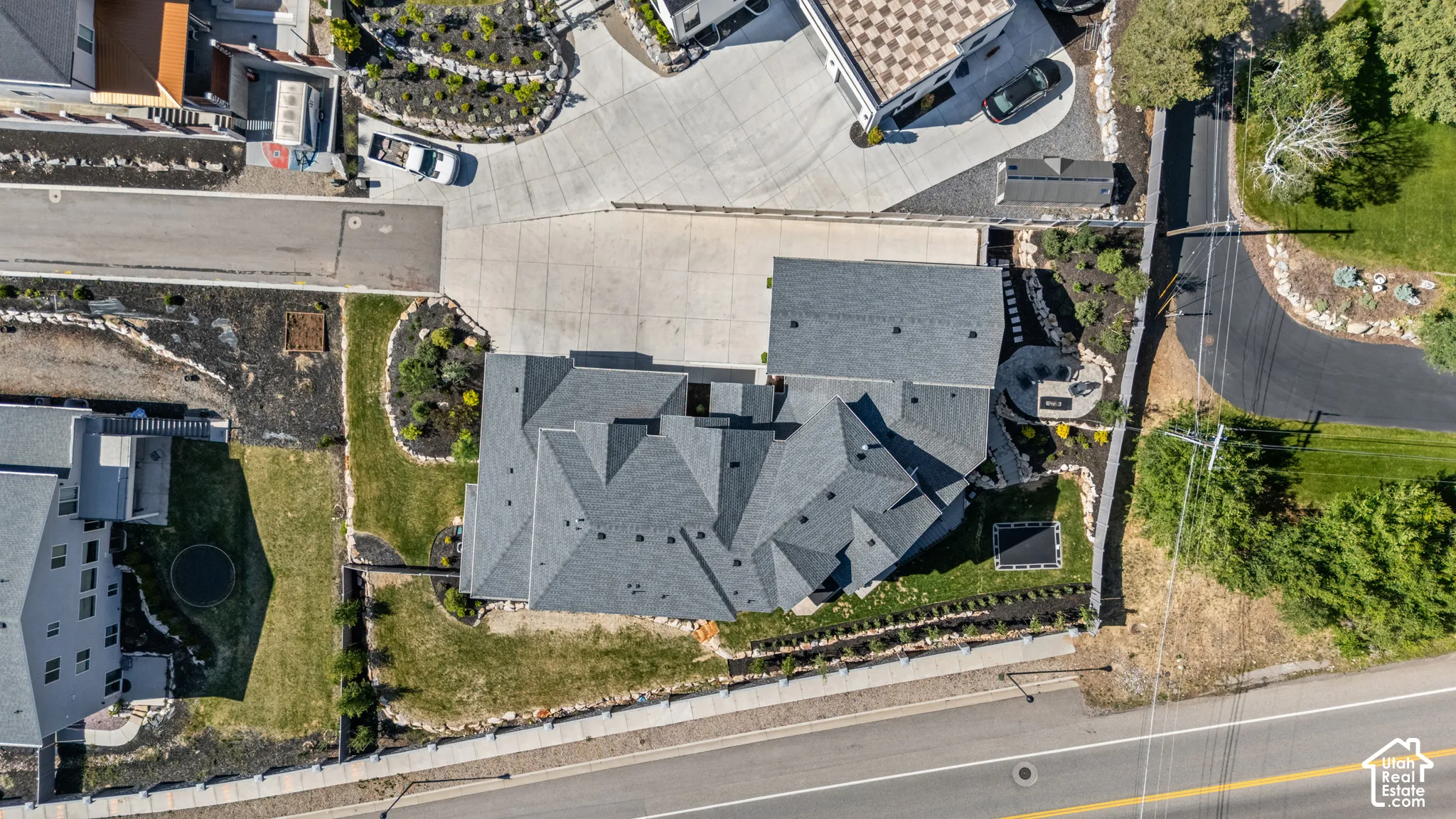 Contemporary residence featuring a multi-gabled gray shingle roof, extensive concrete driveway, landscaped grounds with established plantings, and a stone retaining wall