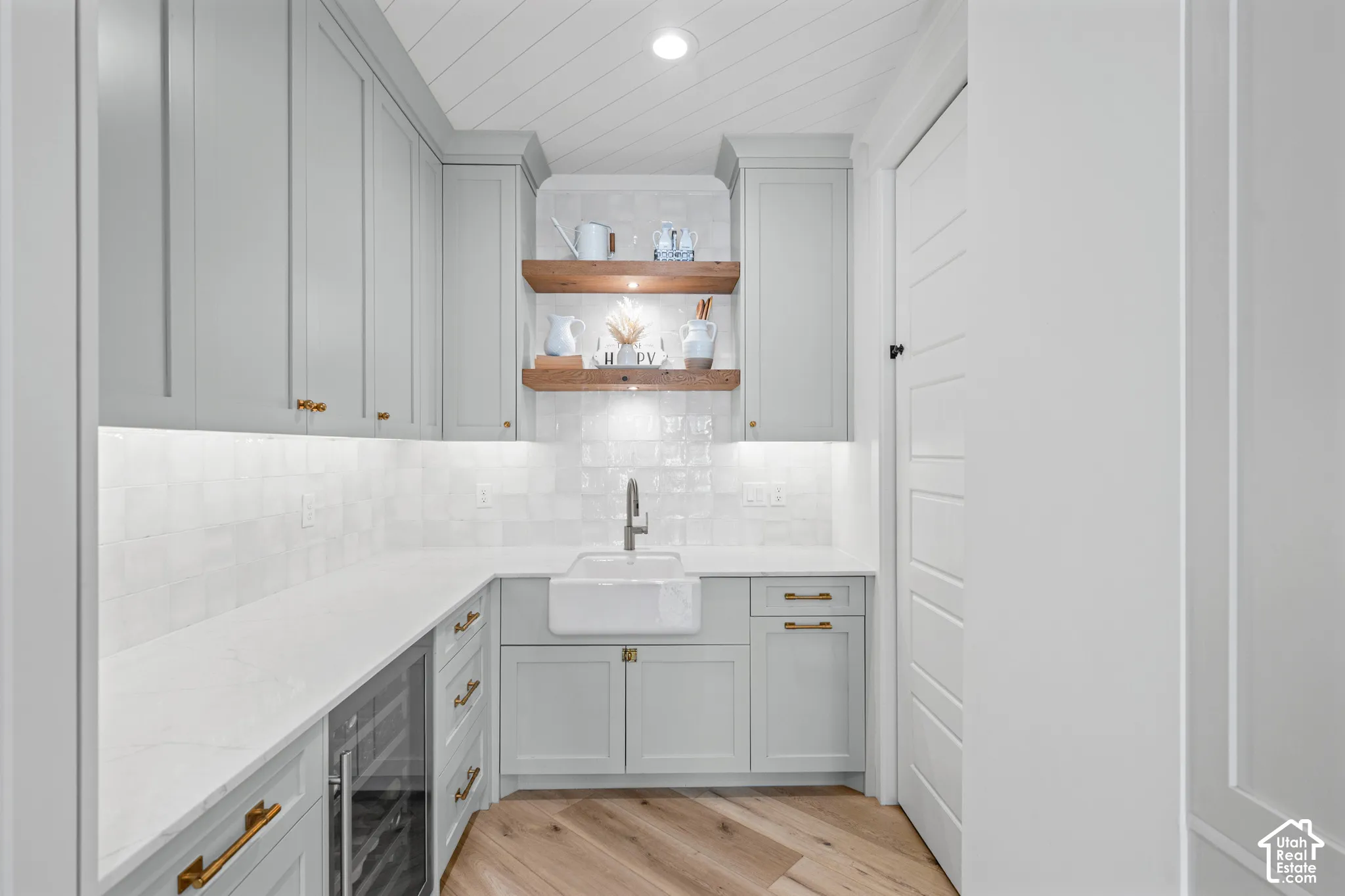 Light grey cabinetry with gold-tone hardware, white subway tile backsplash, white farmhouse sink, and wood-finish flooring
