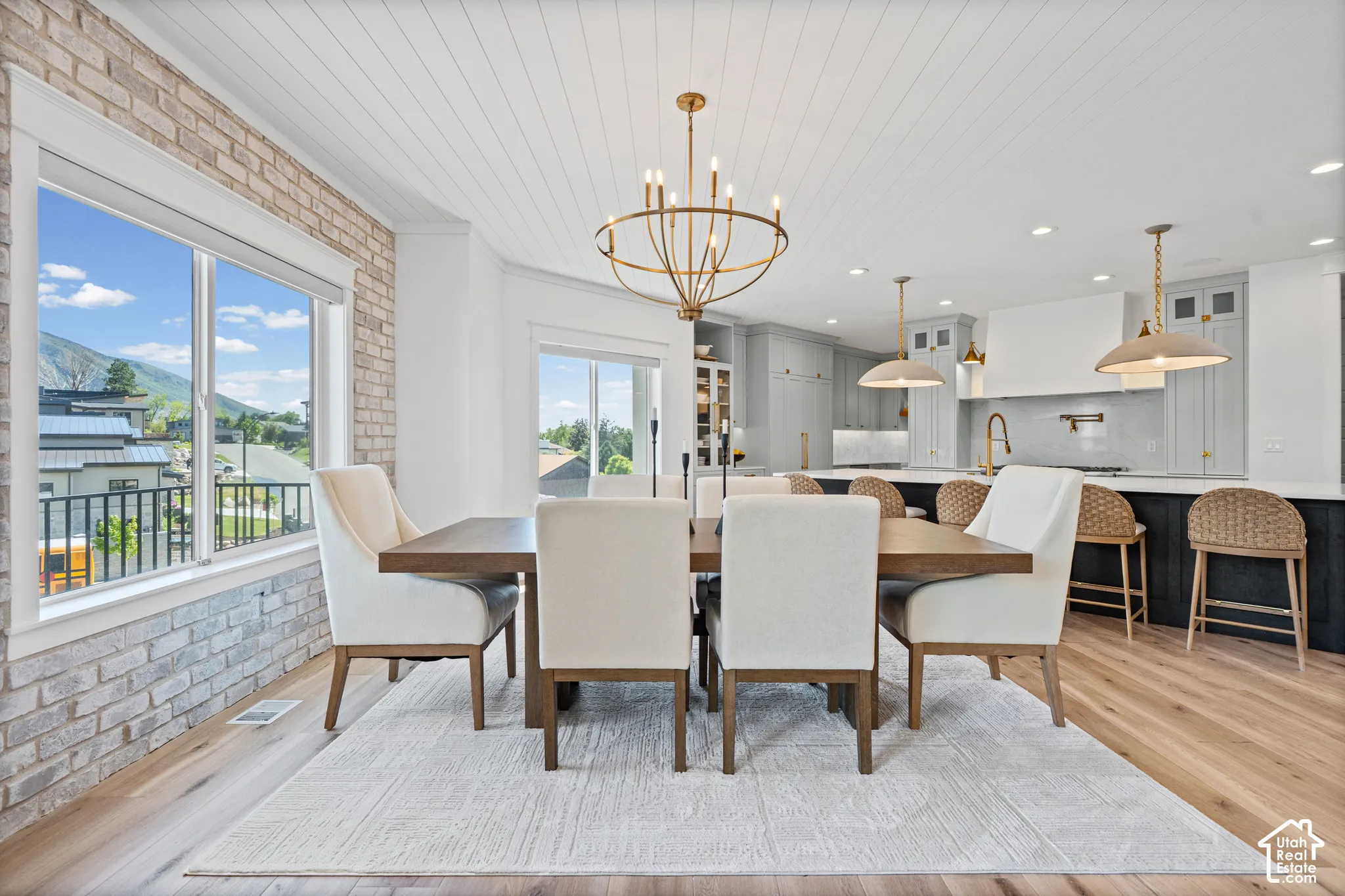 Open-concept dining and kitchen space featuring a large window, wood-finish flooring, a white shiplap ceiling, a contemporary brass chandelier, and a kitchen island with pendant lighting