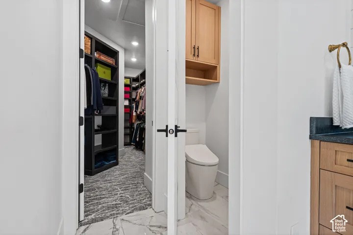 Walk-in closet with dark built-in shelving, patterned flooring, and recessed lighting, adjacent to a water closet featuring a toilet and overhead wood-finish cabinetry