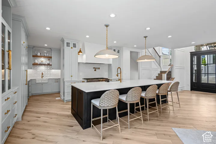 Gourmet kitchen featuring a large central island with seating, light wood-finish flooring, custom cabinetry with brass hardware, and a tiled backsplash