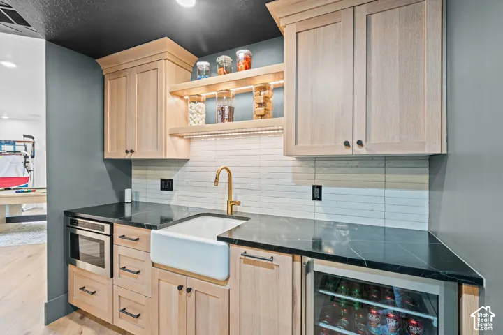 Custom wet bar featuring light wood-finish cabinetry, a white farmhouse sink with a gold-tone faucet, black countertops with subtle veining, and a built-in beverage refrigerator