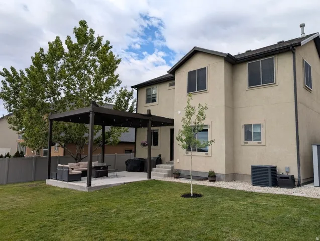 Backyard featuring a concrete patio with a dark-toned pergola, expansive green lawn, mature trees, and a stucco exterior