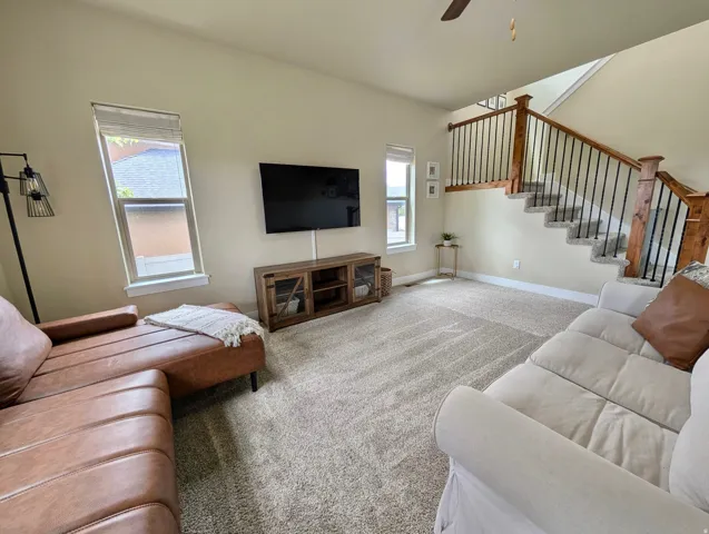 Carpeted living area featuring a wood staircase with black iron balusters, multiple windows, and a ceiling fan