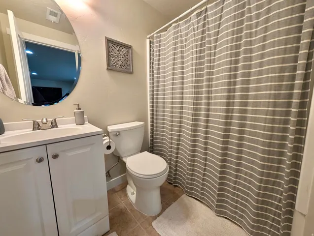 Bathroom featuring a white vanity with panel cabinetry and a polished chrome faucet, a wall-mounted circular mirror, a standard toilet, and tiled flooring