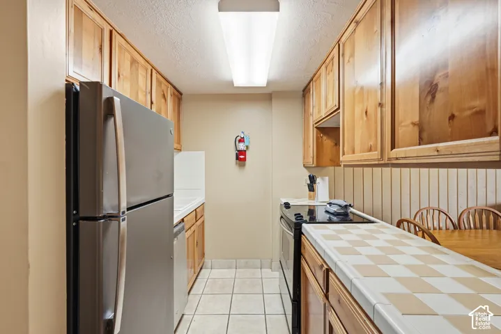 Galley kitchen featuring wood cabinetry, stainless steel refrigerator, tile flooring, and a tiled countertop with a checkered pattern