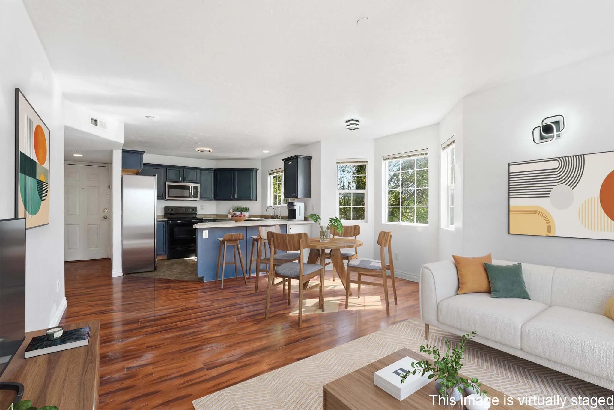 Open-concept living area featuring rich wood-finish flooring, a kitchen with blue cabinetry and stainless steel appliances, a dining area with multiple windows, and recessed lighting