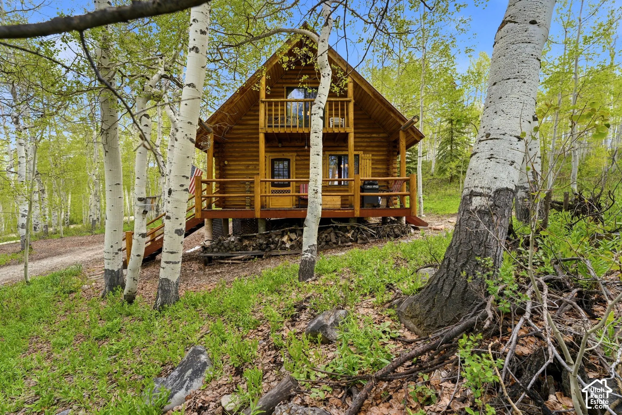 Log cabin exterior featuring a natural wood finish, a covered lower porch, an upper-level balcony, a gabled roofline, and surrounding mature trees