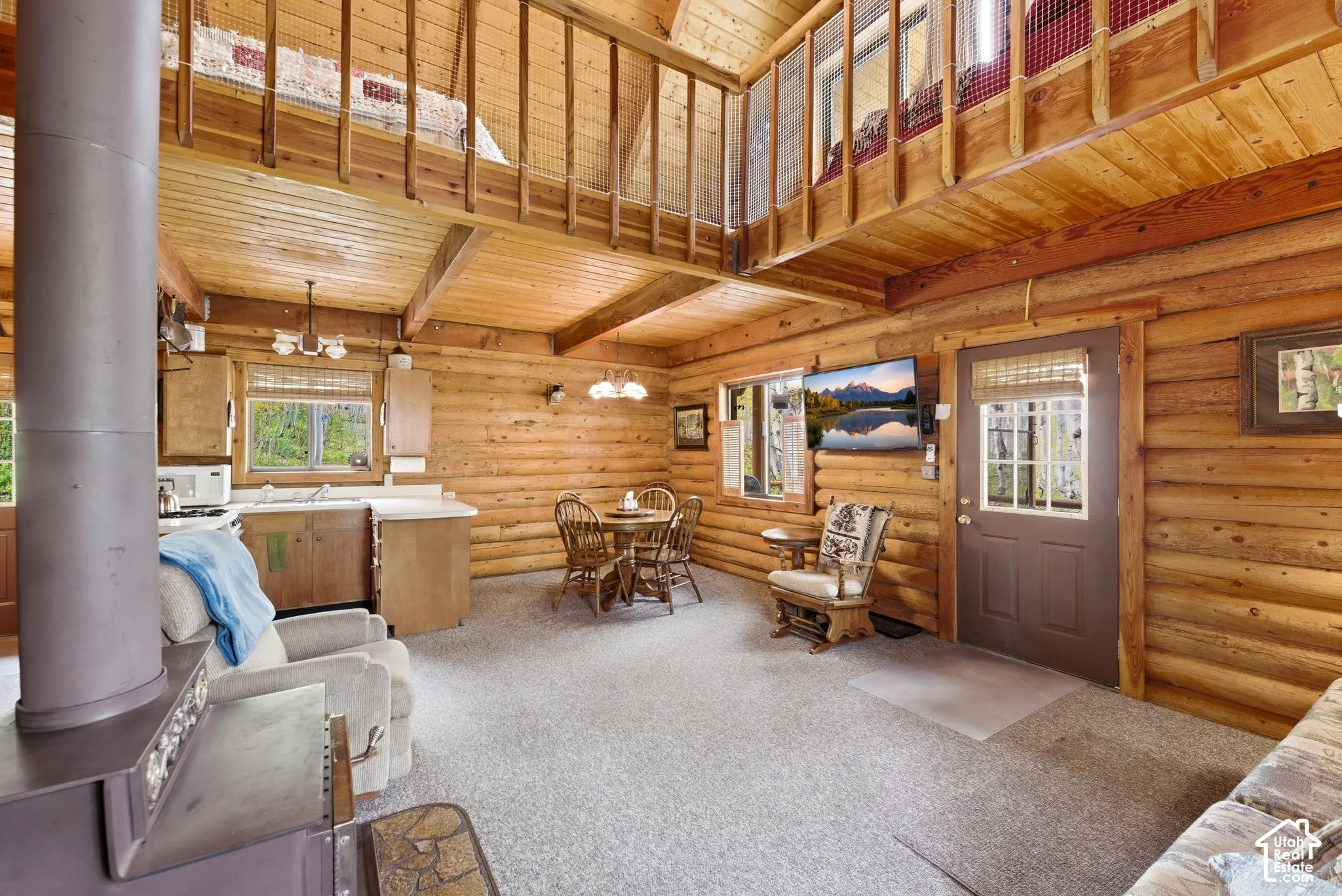 Open-concept living space featuring wood-finish walls and ceilings, a lofted area with wood railing, a kitchen with light wood cabinetry, a dining area with a round wood table, and a built-in wood-burning stove