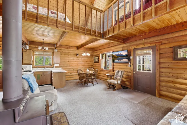 Open-concept living space featuring wood-finish walls and ceilings, a lofted area with wood railing, a kitchen with light wood cabinetry, a dining area with a round wood table, and a built-in wood-burning stove