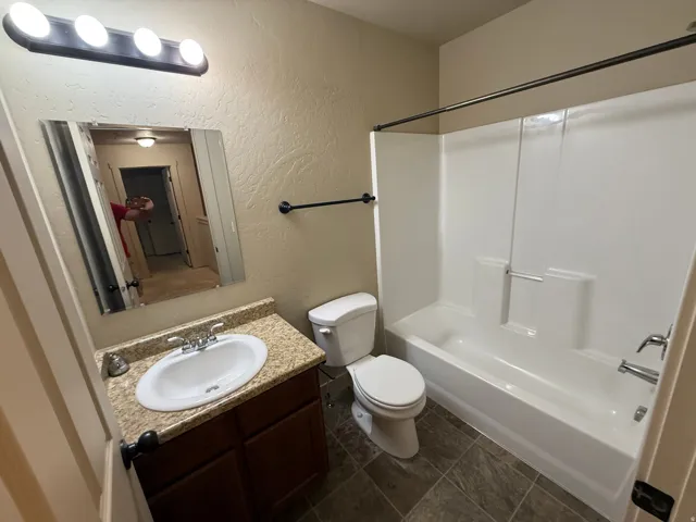Unit B - Bathroom featuring a white tub/shower combination, dark wood-finish vanity with an oval sink, speckled countertop, and tiled flooring