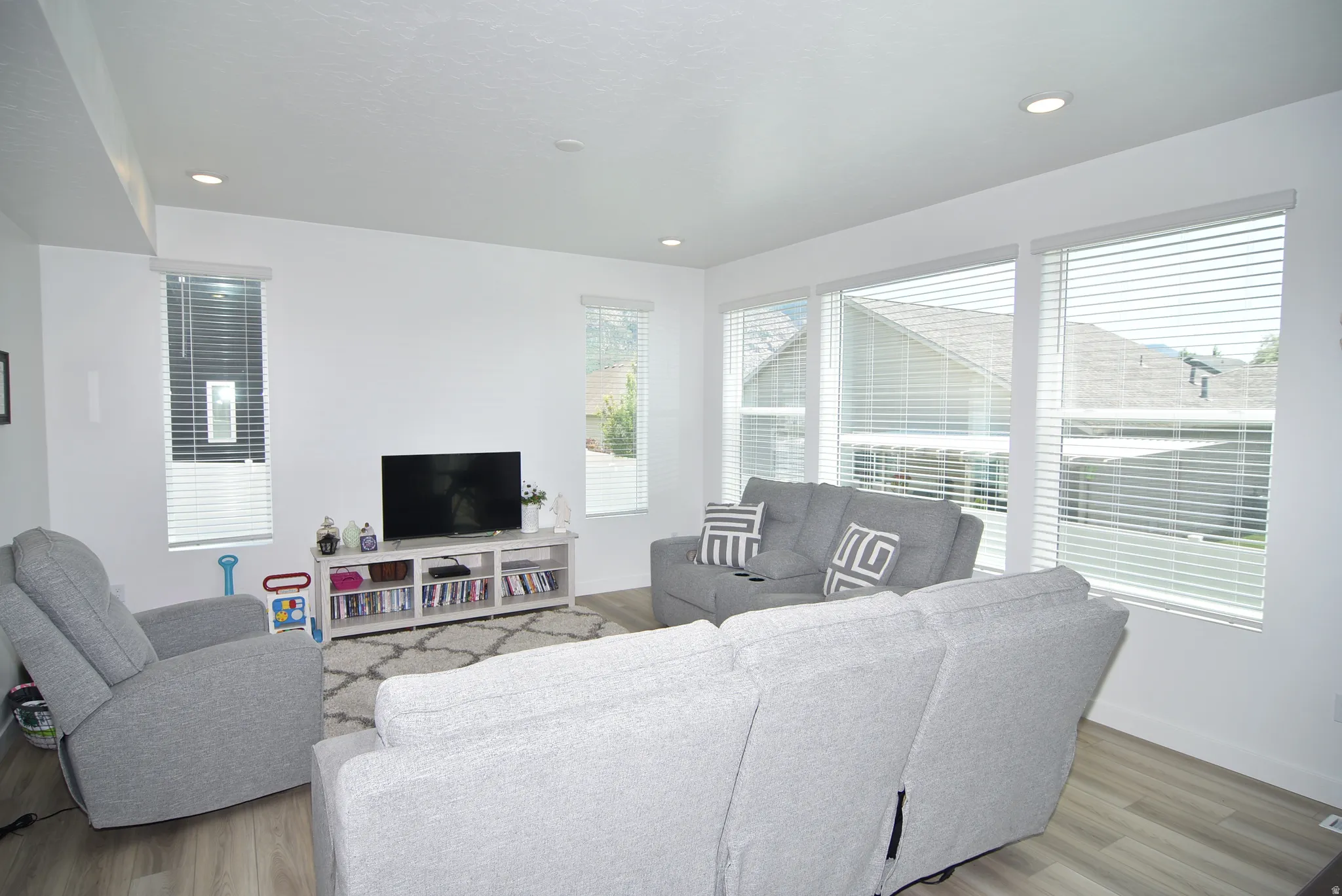 Spacious living area featuring light wood-finish flooring, recessed lighting, and multiple windows with blinds