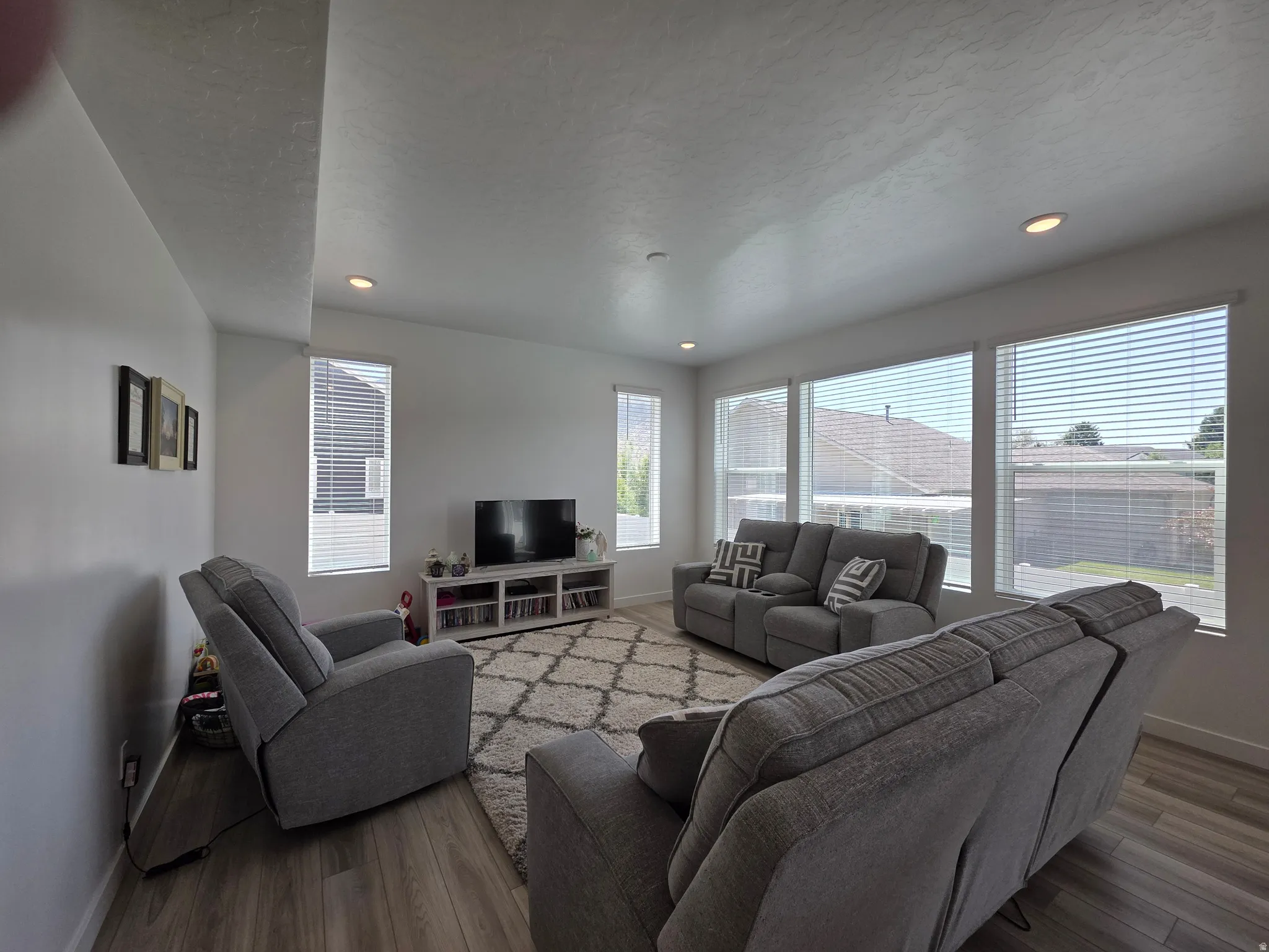 Spacious living area featuring abundant natural light from multiple windows, recessed ceiling lighting, wood-finish flooring, and a media console with open shelving